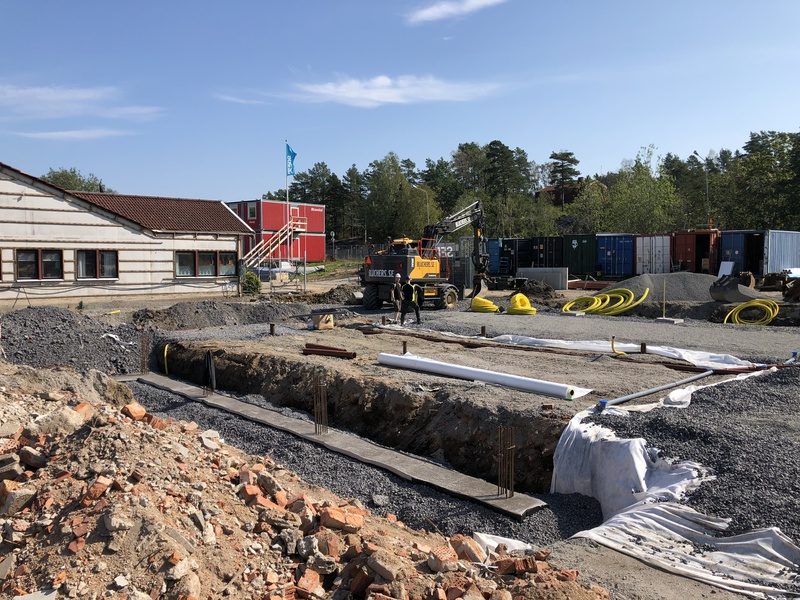 Schakt med grusbädd framför en skolbyggnad, med grävmaskin och etableringsbodar i bakgrunden.